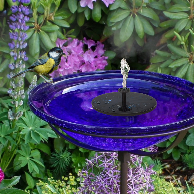 A close-up of a cobalt glass birdbath with solar-powered fountain, perched in a lush flower-filled garden with a bird nearby, perfect for a bird-friendly garden.

